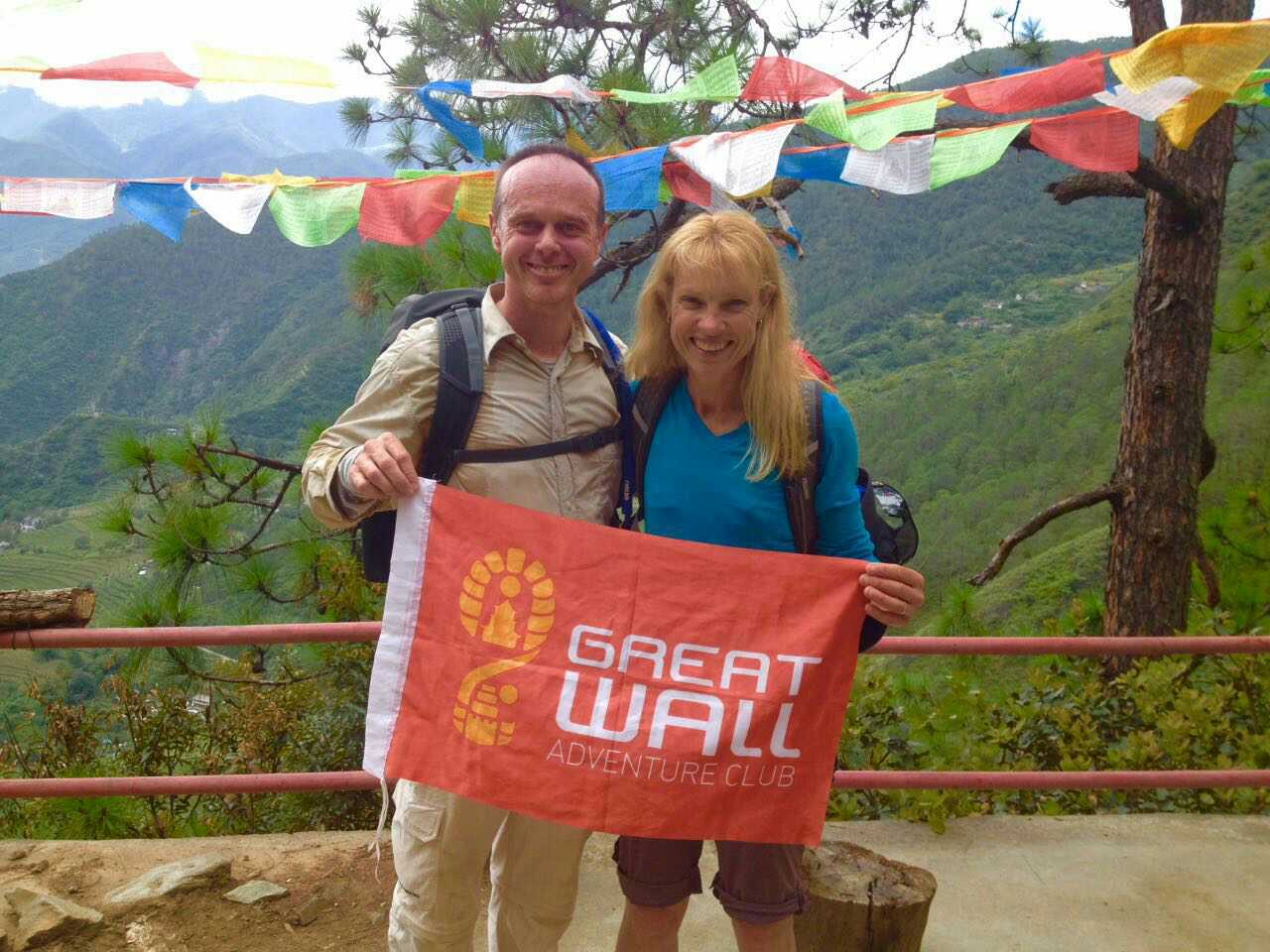 Two travelers holding a Great Wall Adventure Club banner beneath colorful prayer flags